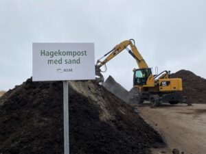 En gravemaskin graver i en av flere store komposthauger på Årabrot miljøpark. Et skilt viser at det er hagekompost med sand det graves i.