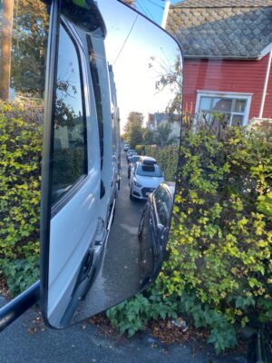 Bildet viser speilet på en renovasjonsbil i en trang gate, der det er høye hekker og biler parkert langs veien.
