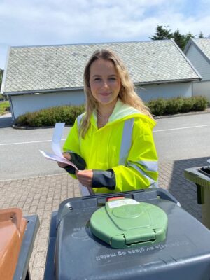 REdu-student Silje Bie Helgesen står ved en beholder for glass- og metallemballasje. Hun har klistremerker med QR-koder i hendene, og har limt et merke på beholderen.